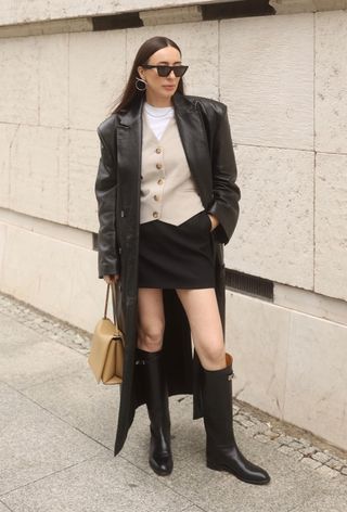 A woman wearing a black leather trench coat styled with a tan vest coat, a white t-shirt, black miniskirt, black knee-high riding boots, and a beige bag.