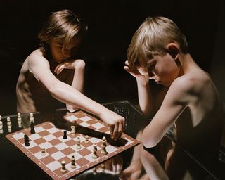 Two boys without shirts on playing chess on a reflective table.
