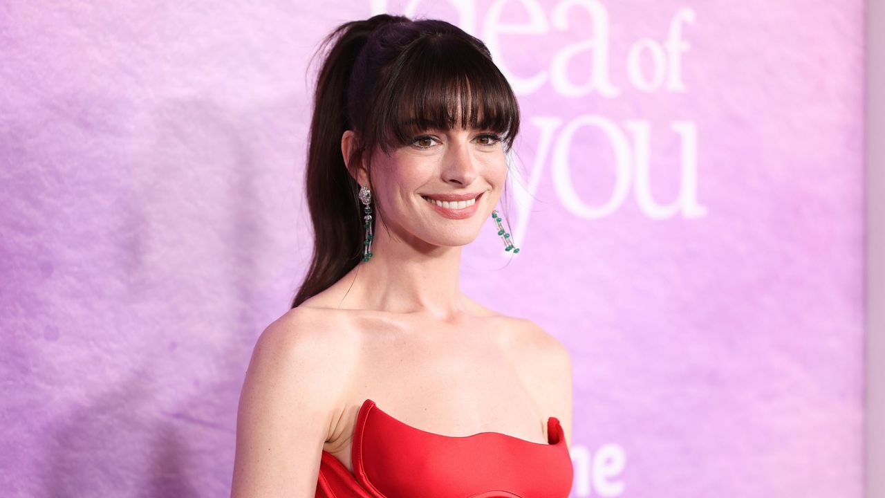 Anne Hathaway&#039;s kitchen is so chic. Here is the actor with a dark brown fringe and ponytail, wearing a strapless red dress, standing in front of a purple background with white writing on it