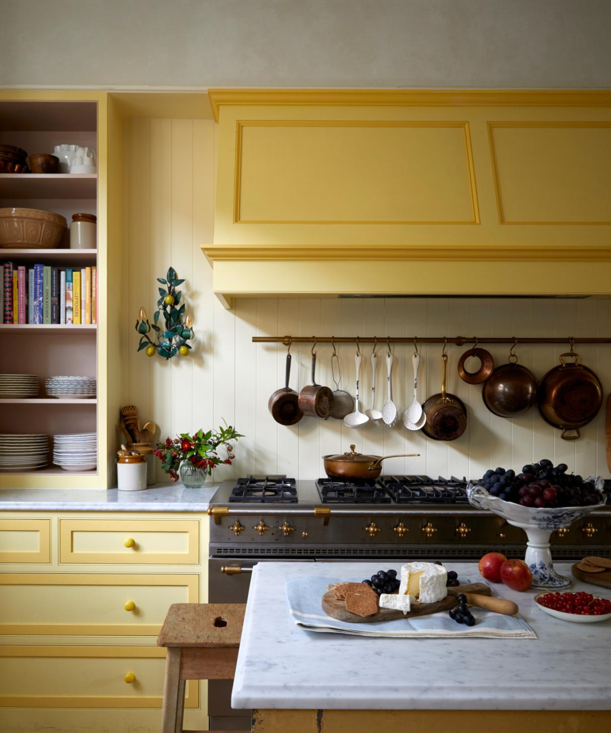 a kitchen with yellow cabinets, a yellow range hood, a line of pots and pans lined up and hanging, and food on the countertop