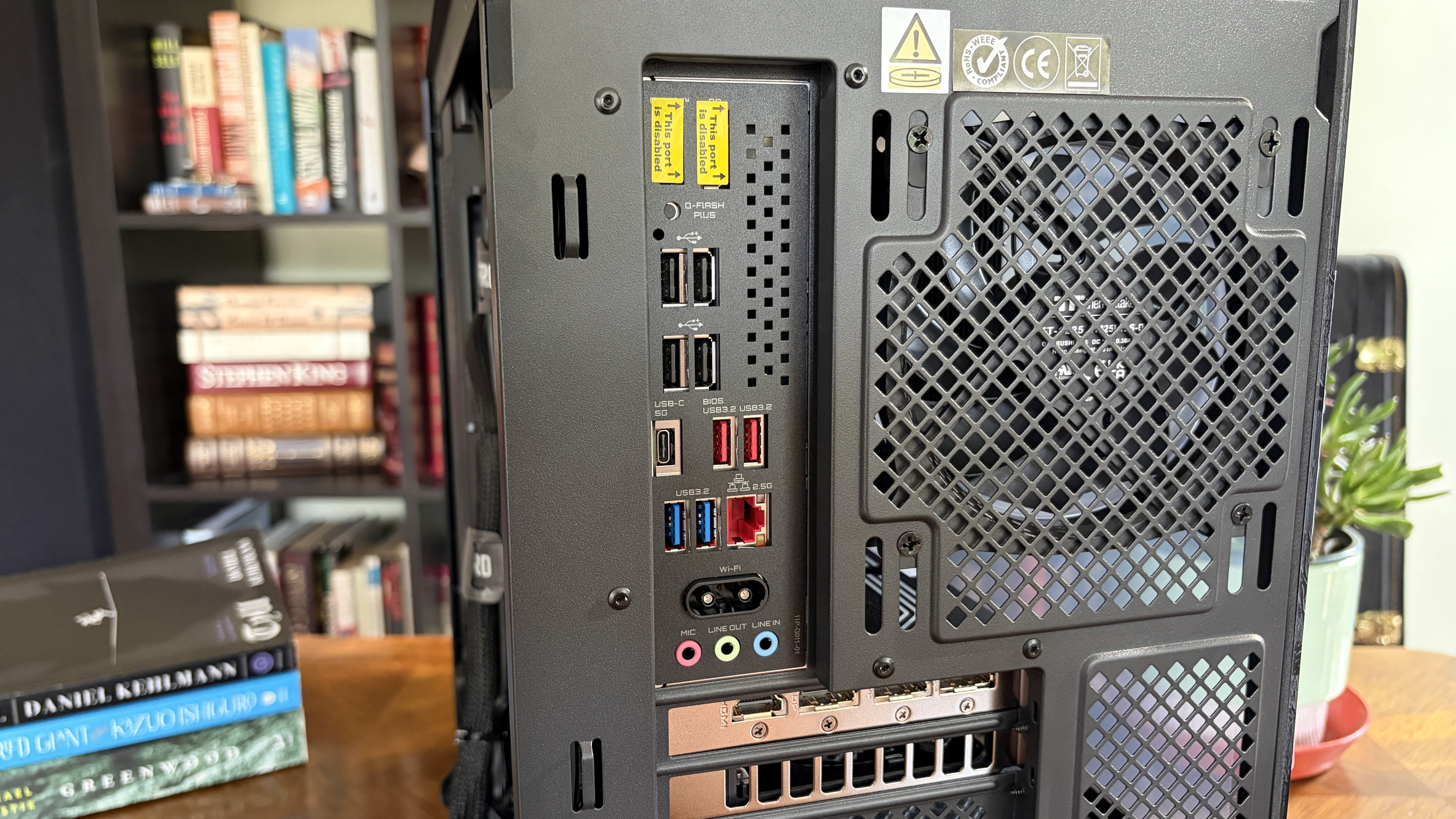 Back view of a Nitro 65 computer tower showing various input and output ports, including USB and HDMI, with a bookshelf and potted plant in the background.