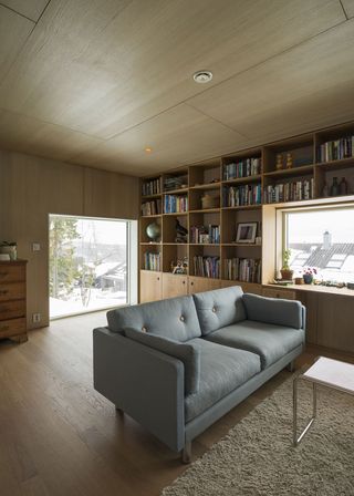 Alternative interior view of a living area at the Oslo family house featuring a wood panelled ceiling with spotlights, wood panelled walls, wood flooring, a blue sofa, an off-white coloured rug, a small table, a wooden shelving unit with books and other items, a chest of drawers and windows