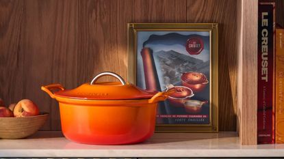 An orange Dutch oven on a wooden shelf, beside a framed illustration of the Le Creuset Modern Heritage pan collection, and a wooden bowl of apples.
