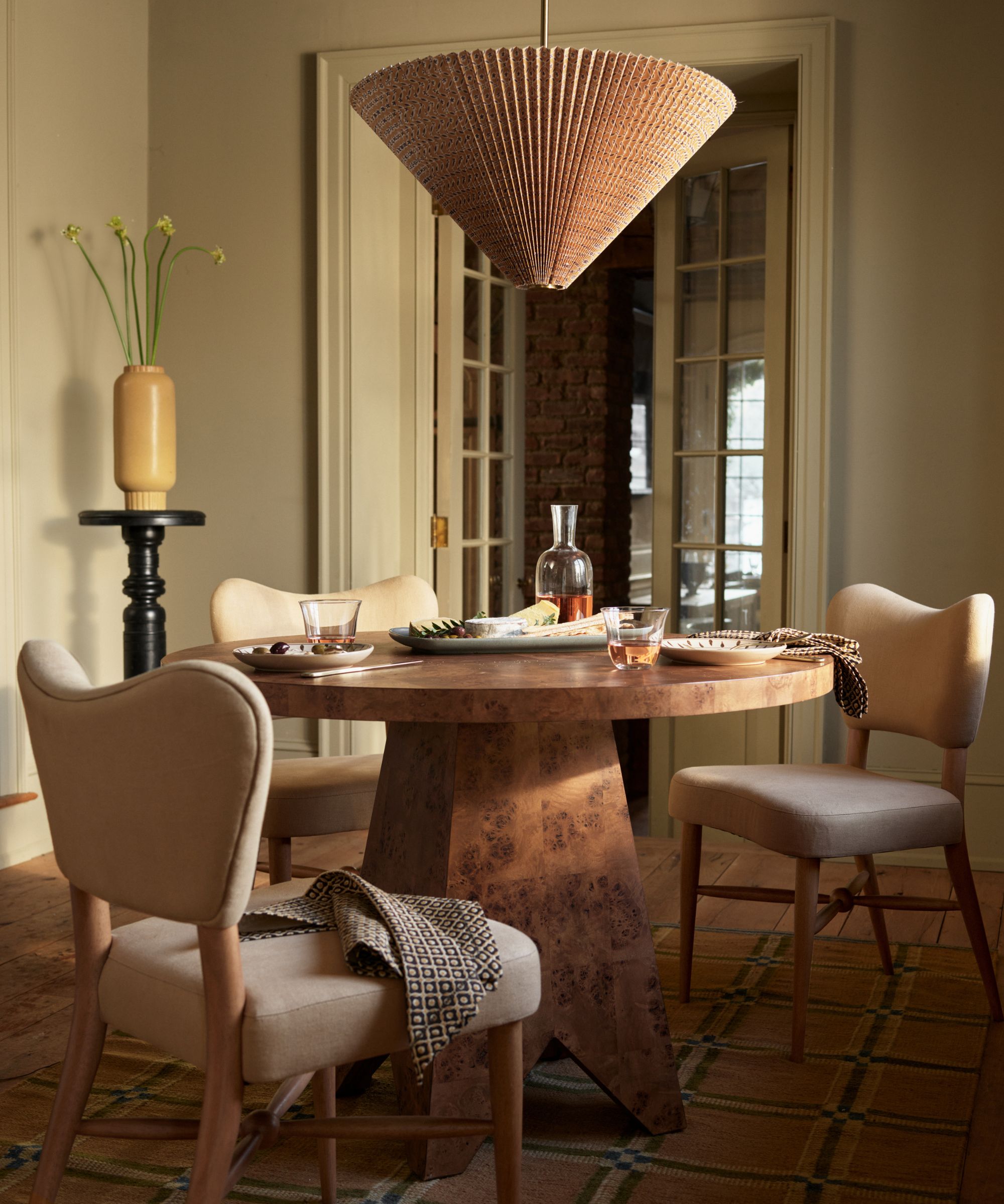 pierce and ward for west elm dining space with a burl wood round table upholstered wood dining chairs and a pleated ceiling light above