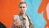 Portrait of teenage girl in glasses with short hair in leopard jacket using mobile phone against city street wall