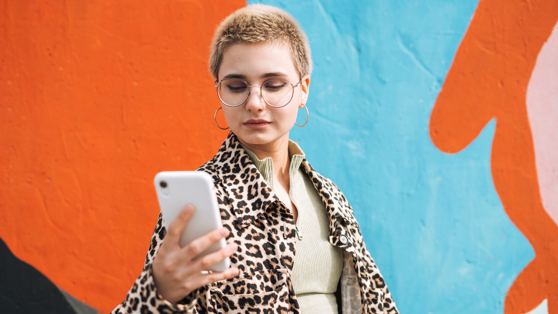 Portrait of teenage girl in glasses with short hair in leopard jacket using mobile phone against city street wall