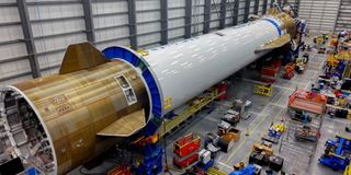 A long fuselage lays on the left on the floor of a large factory..