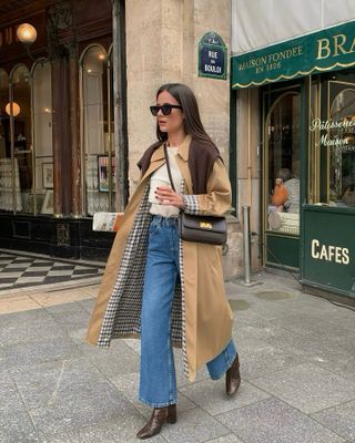 Woman wearing white shirt, blue jeans, and trench coat, walking in front of Paris cafe.