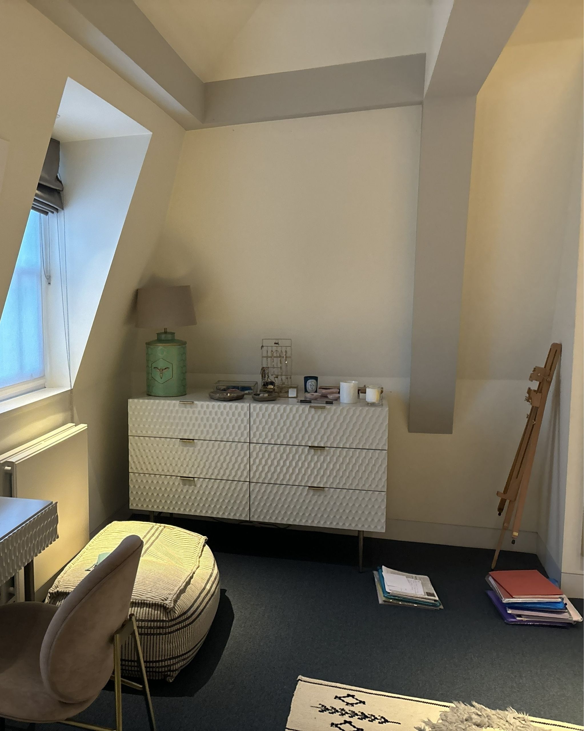 The corner of a white room with navy carpets. There is a white dresser with a green lamp on it, a wooden easel, and a window. 