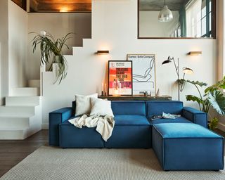 A teal blue L-shaped modular sofa in cream living room with wall art, pair of wall sconces and staircase in background
