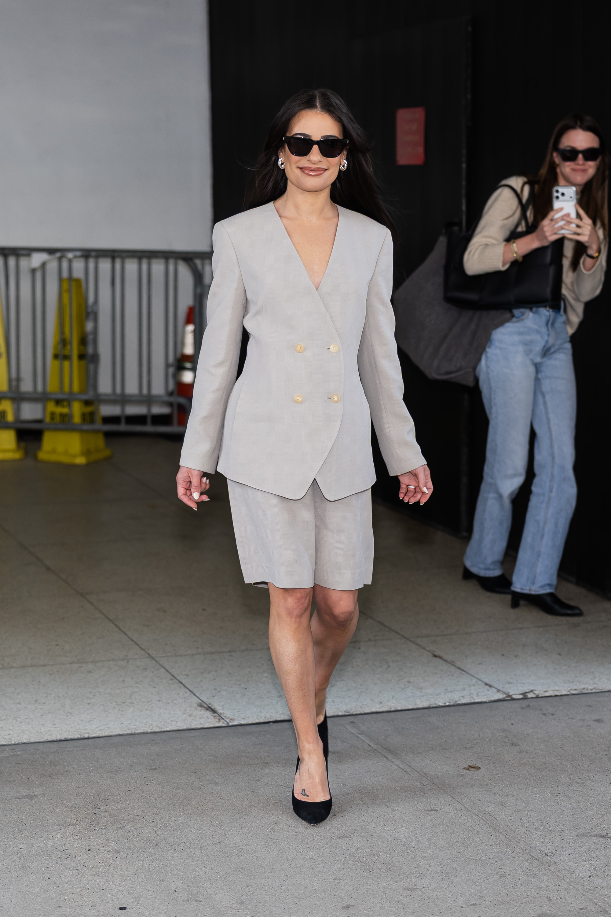 Lea Michele wears a gray blazer with gray Bermuda shorts and black pumps.