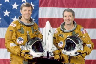 two smiling men in orange-brown flight suits stand in front of an american flag