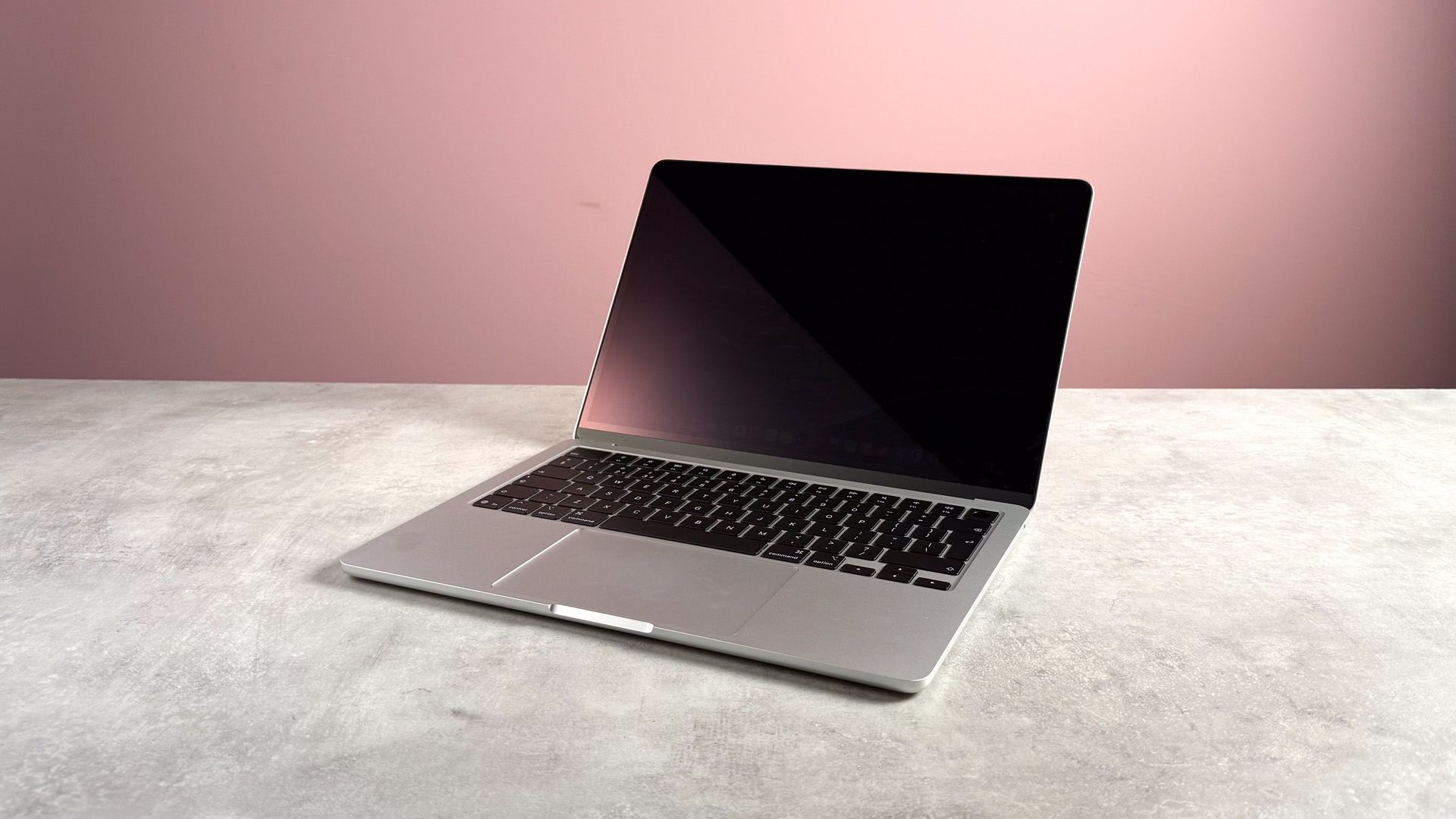 The MacBook Air M5 open 120 degrees but powered off on a stone effect surface in front of a pink background.