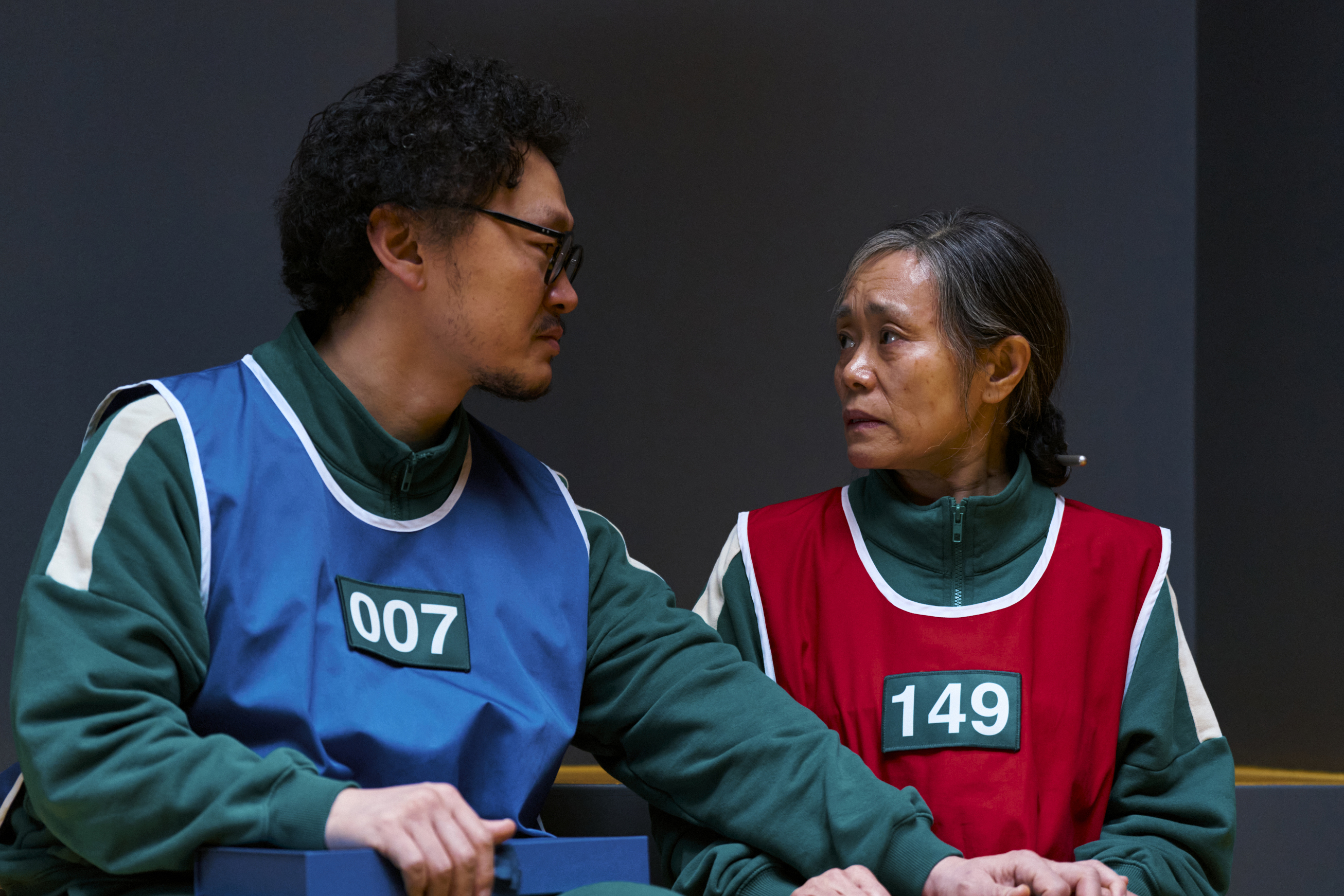 A man (Yang Dong-geun as Yong-sik, Player 007) holds the hand of his elderly mother (Kang Ae-shim as Geum-ja, Player 149) as they wear different colored vests, in 'Squid Game' season 3.