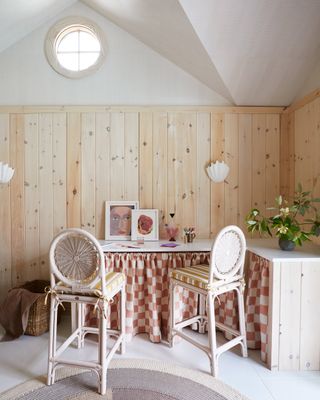 Image of an office that is white with light wooden-paneled walls. The desk is a corner desk with a terracotta and white checkered skirting covering the front. There are two round, wicker stools, and paintings and a plant on the the desk.