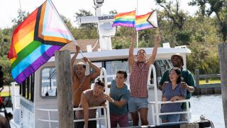 From left to right: Tomas Matos waving a giant pride flag, Joel Kim Booster leaning over the edge of a boat, Conrad Ricamora smiling, Matt Rogers holding up and waving two pride flags, Margaret Cho and Torian Miller standing together smiling in Fire Island.