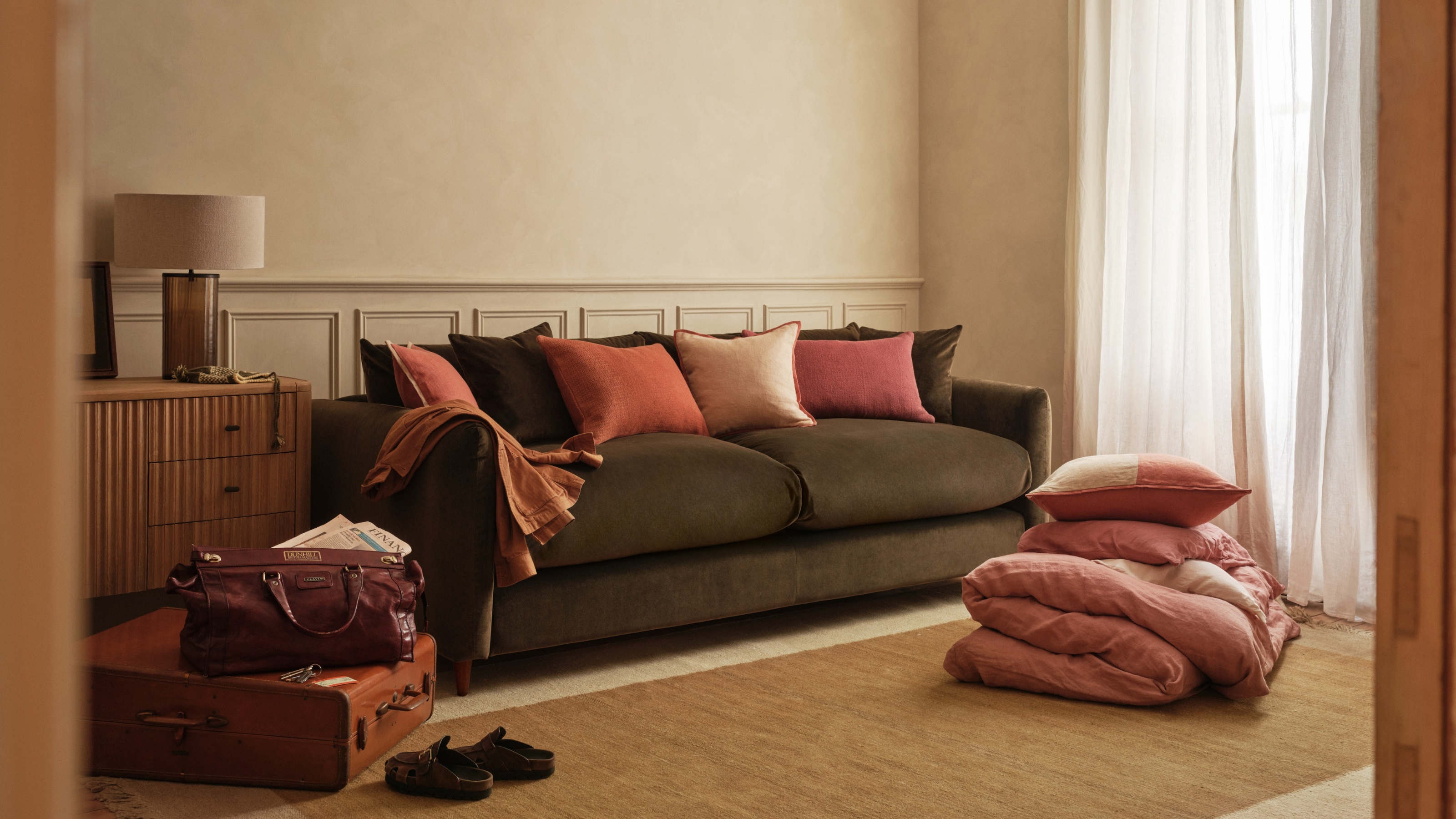 Image of a dark green loaf sofa bed with pink pillow in a warm, beige living room. There is a jute rug on the ground with pink bedding folded on it, and a suitcase in the corner.