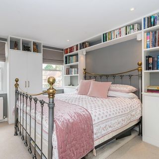 Bed with white and pink bedding and silver and brass headboard and footboard on beige carpet in front of grey wall with white bookshelves packed with books