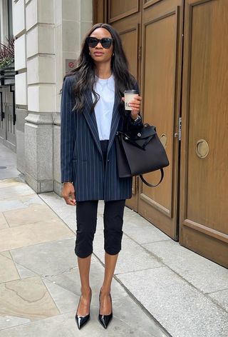 a woman wearing black capri pants with a white t-shirt, navy pinstripe blazer, black bag, and black pointed pumps