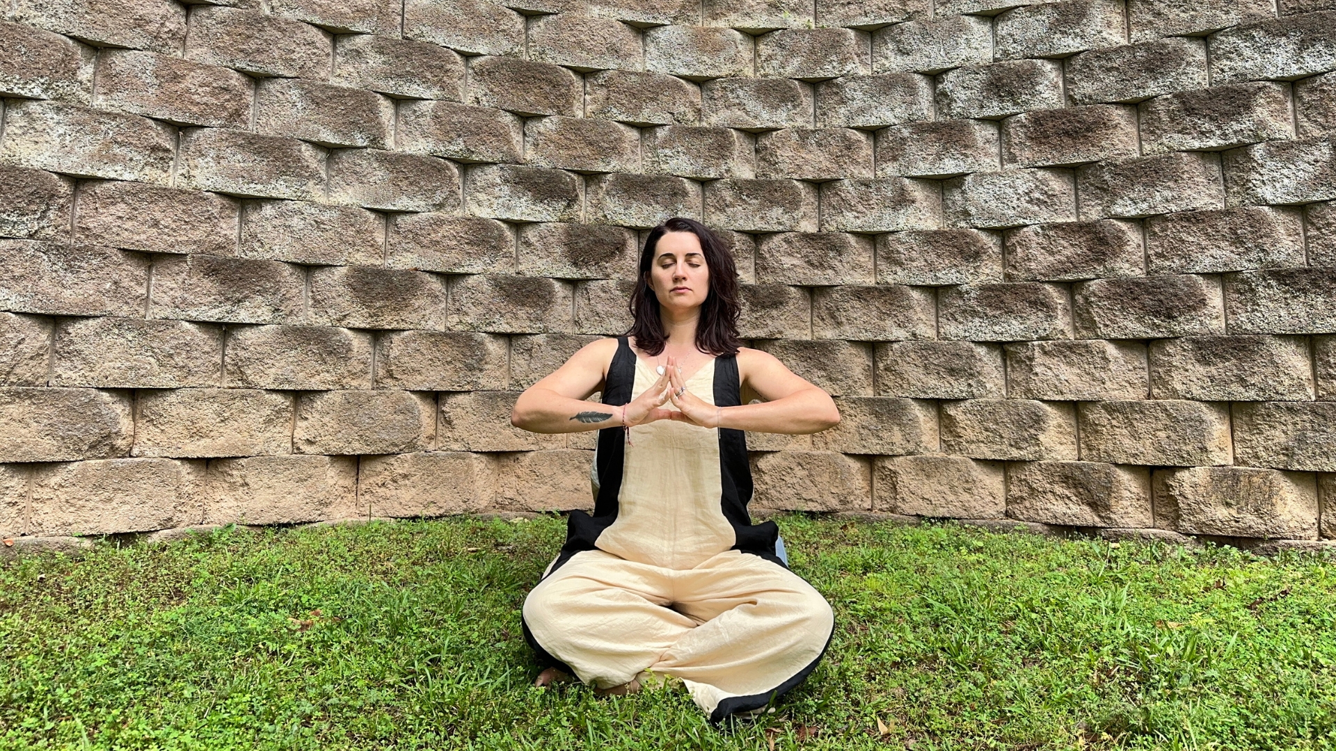 woman sat cross legged with fingers touching in front of her chest. she's on some grass with a large stone wall behind her.