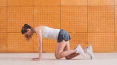 woman wearing a white vest and grey shorts performing a bear crawl exercise in front of an orange tiled wall