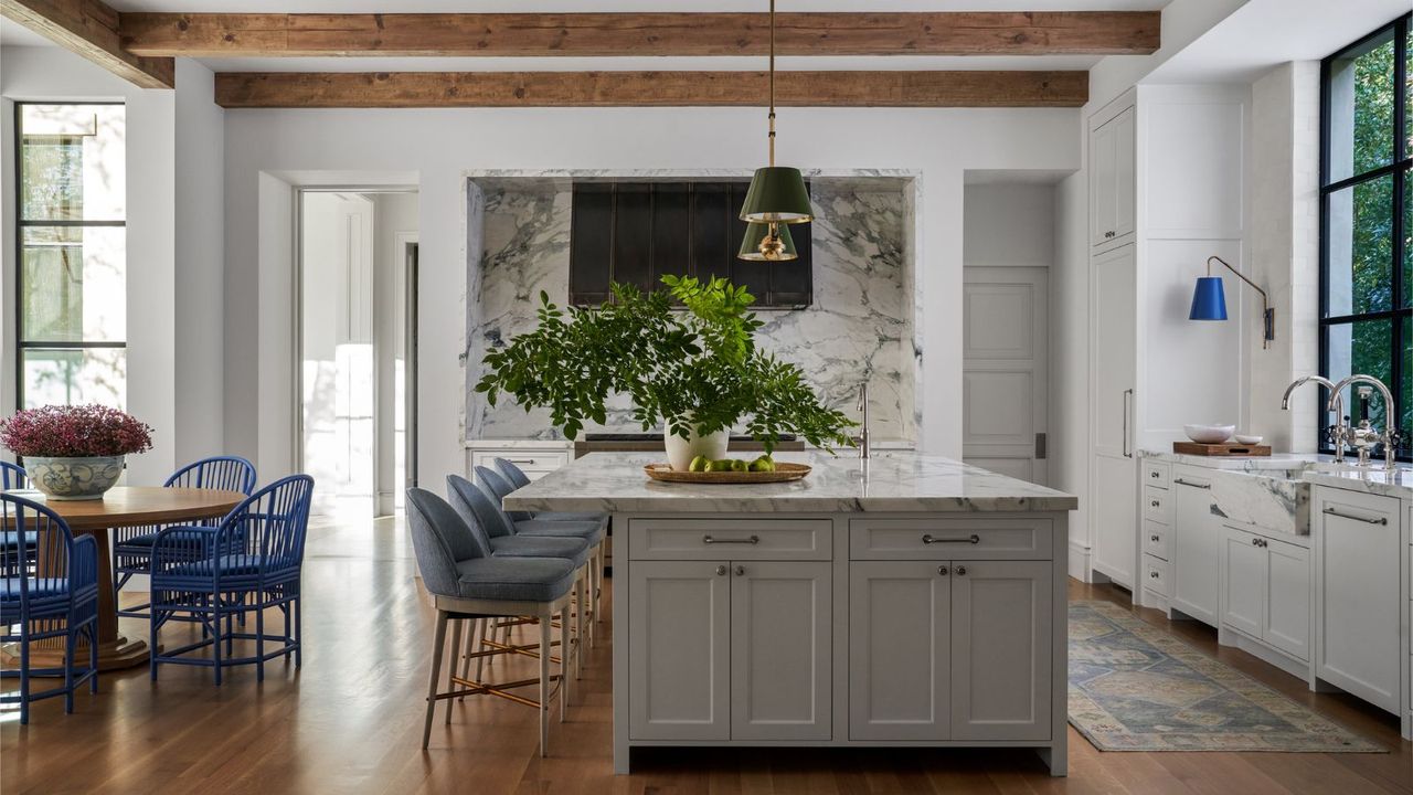 a great room with a kitchen and a dining space. On the kitchen counter is a large plant and four barstools are positioned to the left. To the left is a round dining chair with four blue chairs. Pendants hang from a ceiling with exposed beams