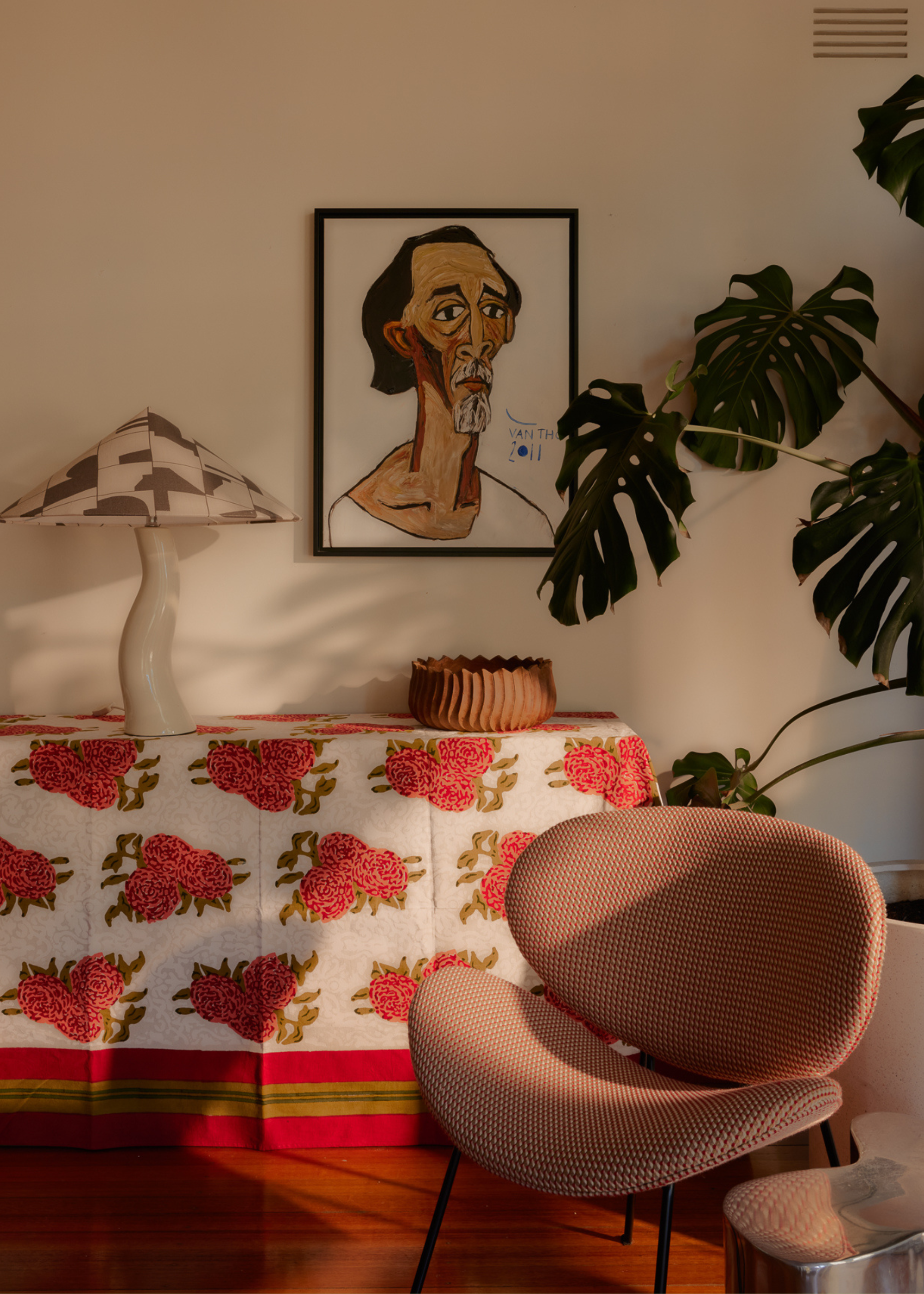 A monstera in a living room with a patterned chair in front of a credenza with a wavy lamp, a patterned cloth, and a framed piece of art