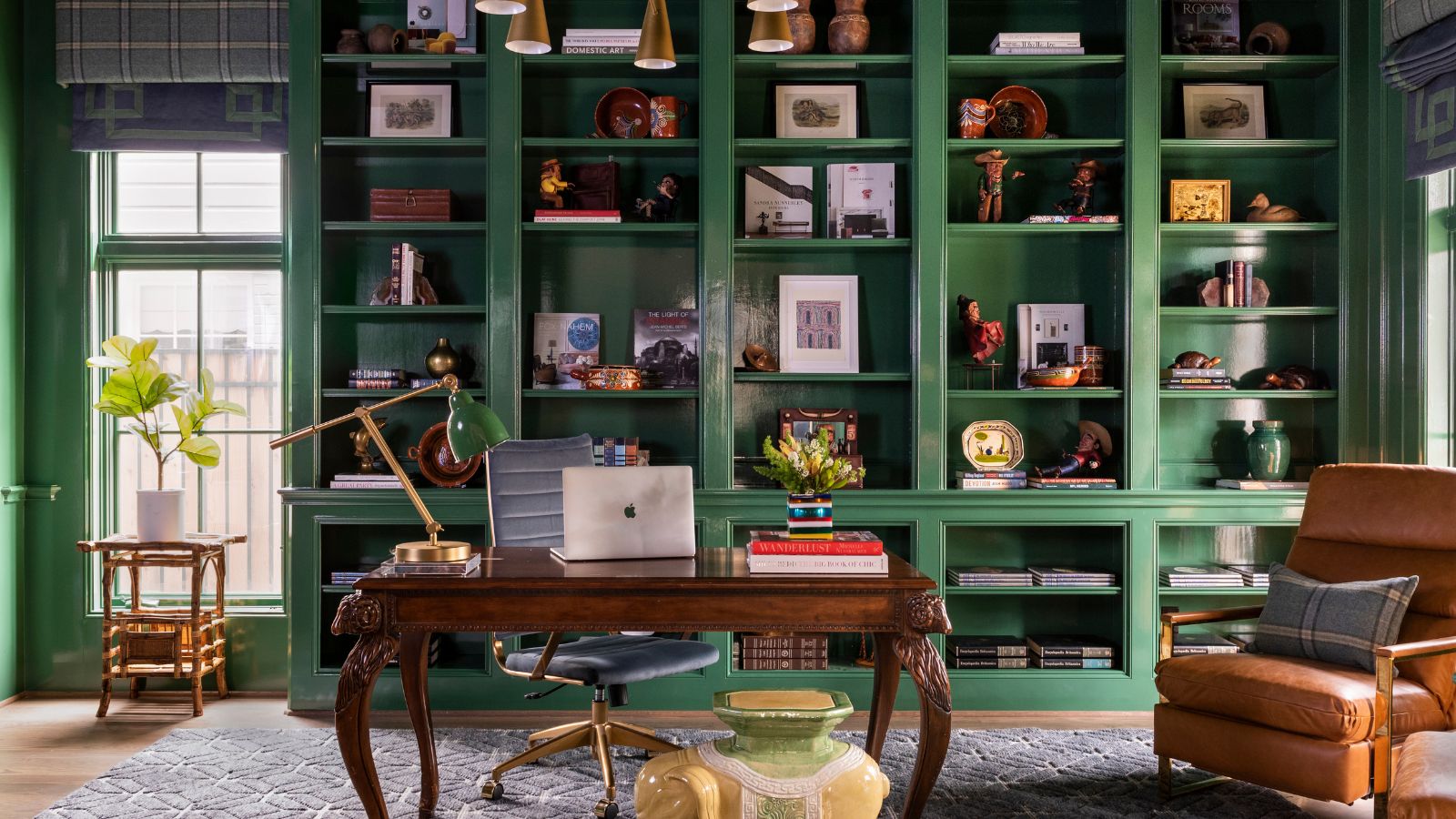 A library room with green gloss paint on all the woodwork and built-ins, eclectic decor on the shelves, an antique wooden desk and a tan armchair.