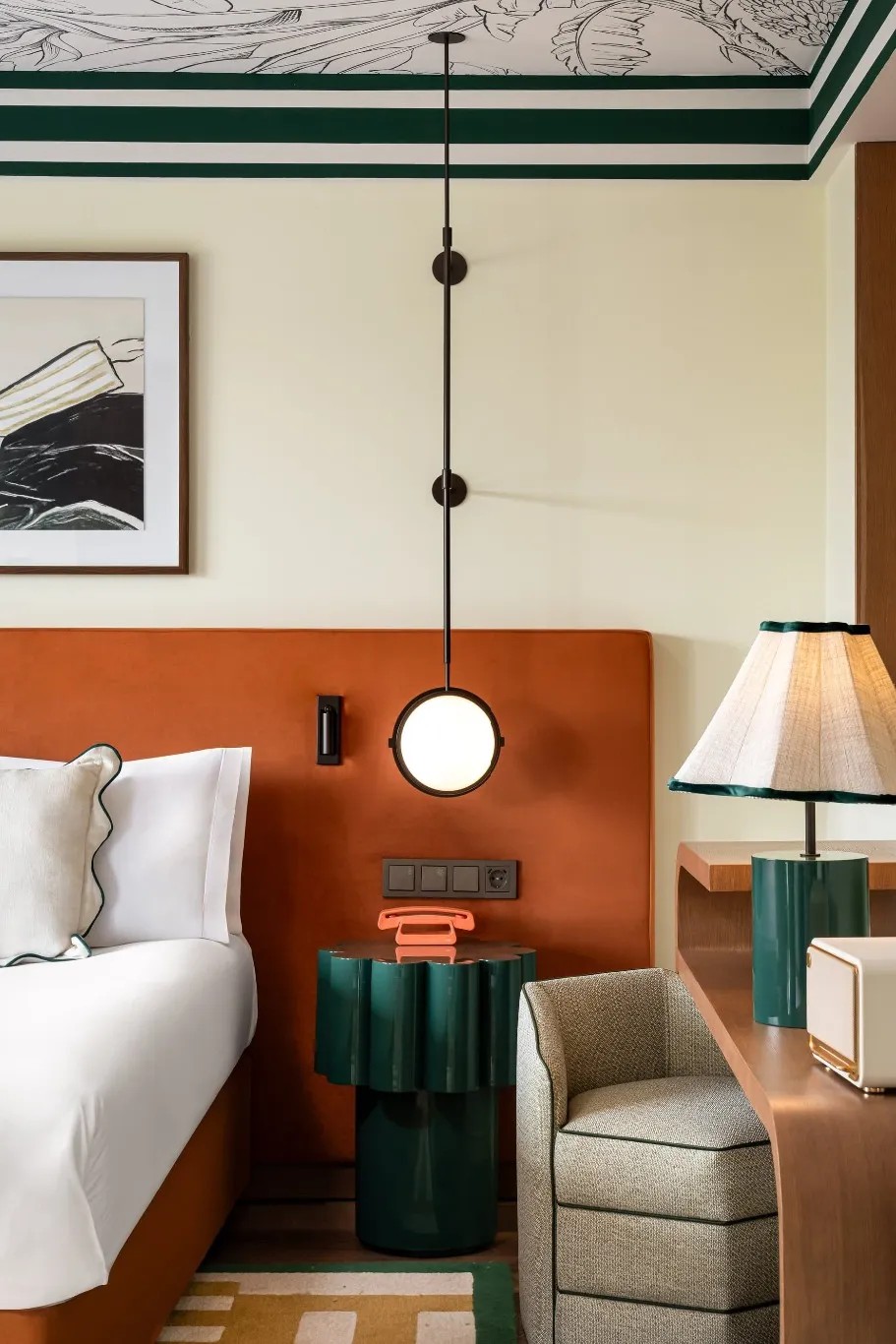 A hotel suite with a black-and-white, eccentric mural, similarly styled artwork, pale yellow walls, dark green and orange accents throughout, mixed with lacquered wood furniture and spherical lights.