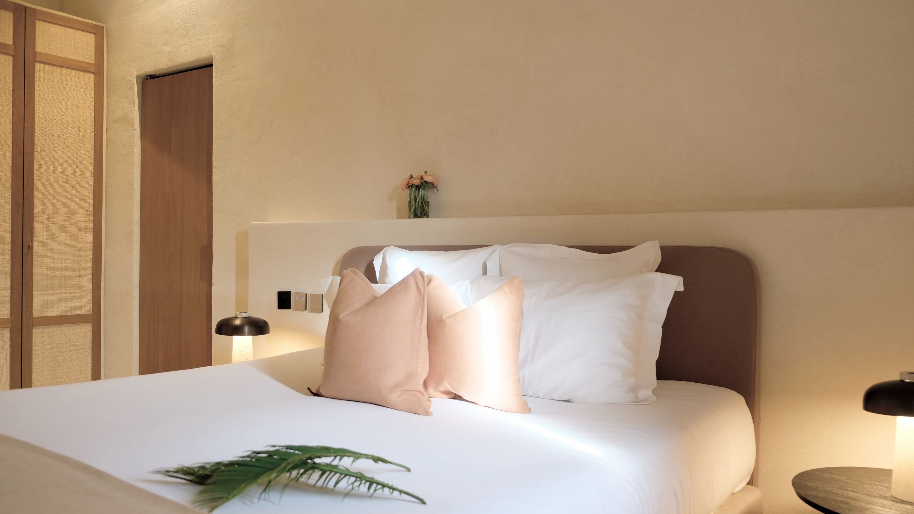 A minimalist hotel bedroom with warm plaster walls, featuring a bed with plush white linens, peach-toned accent pillows, and integrated bedside lighting.