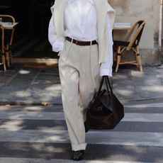 Woman wears cream trousers, a white shirt and carries a brown The Row Margaux leather tote bag