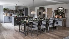 A black, white and grey kitchen with grey cabinetry, and wooden floors. There's a grey kitchen island in the background, and a large laid dining table in front, covered in branches, glasses, lit candles, with black and white blankets on the chairs. To the right is a circular mirror. The walls are white.