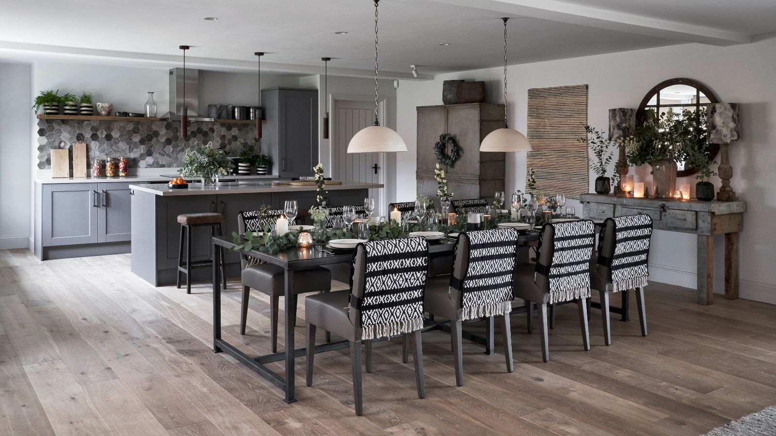 A black, white and grey kitchen with grey cabinetry, and wooden floors. There&#039;s a grey kitchen island in the background, and a large laid dining table in front, covered in branches, glasses, lit candles, with black and white blankets on the chairs. To the right is a circular mirror. The walls are white.