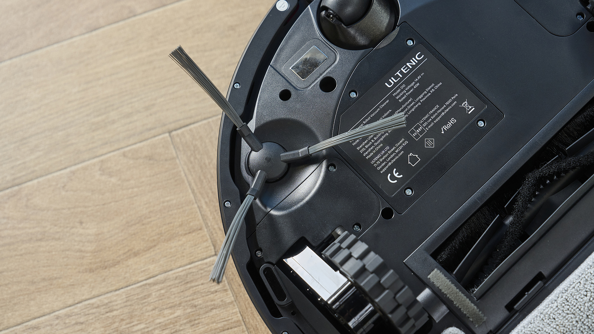 a rechargeable robot vacuum and mop in black by ultenic is photographed on a hardwood floor with a blue wall and a burnt orange terracotta and white striped rug