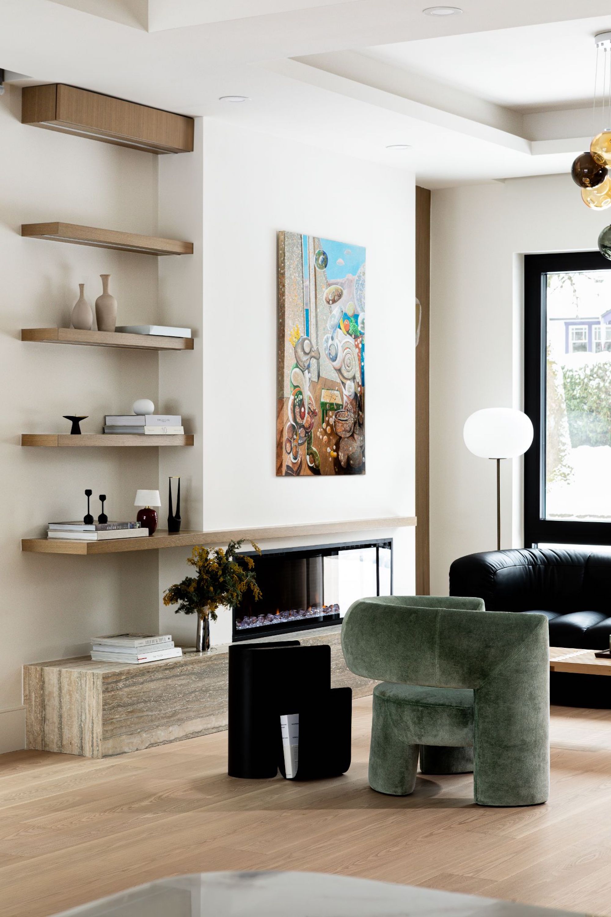 A view of the living space showing the fitted bookshelves and the fireplace with an a surrealist artwork above, as well as the modern, curved armchairs looking outwards to the windows.