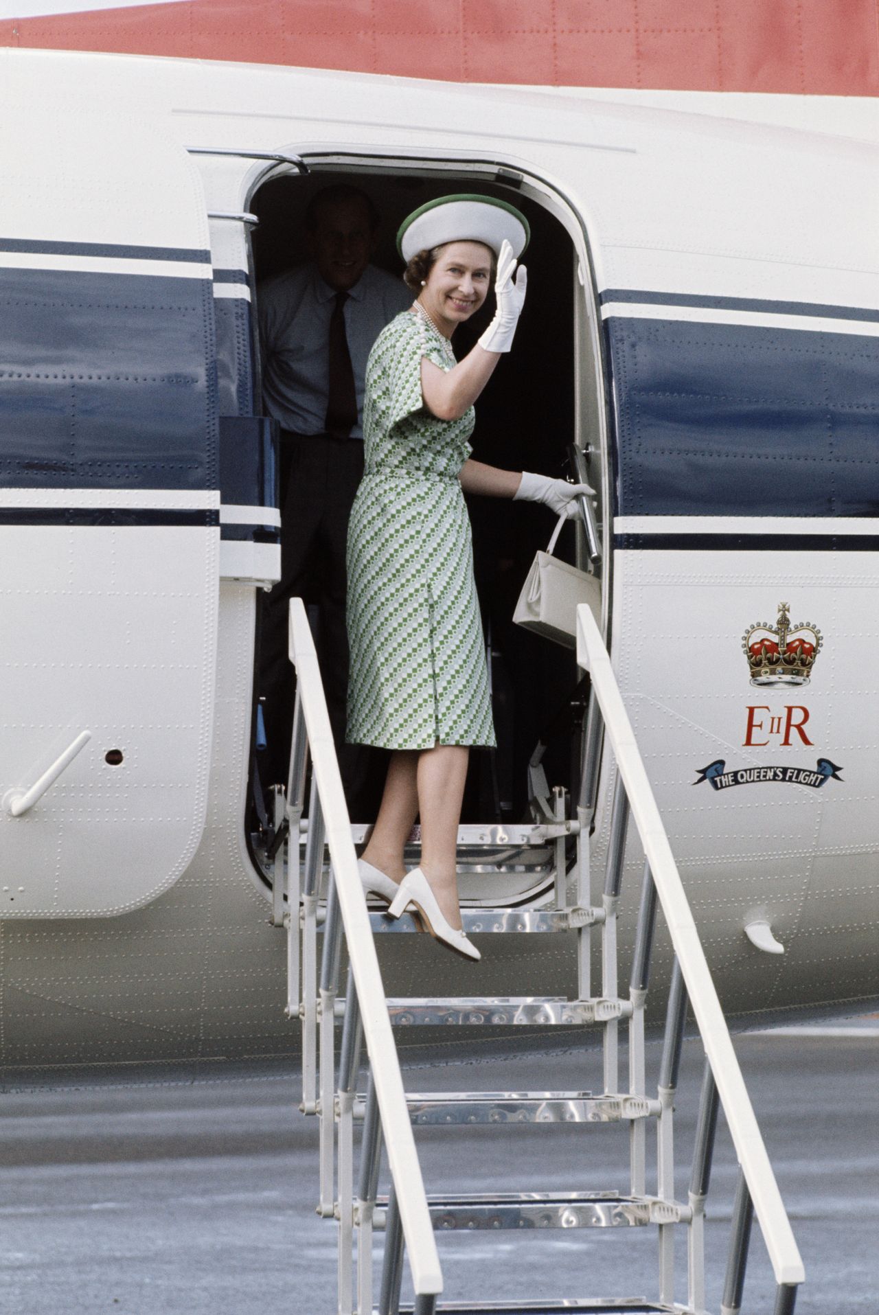 Elizabeth II, wearing a green patterned, short-sleeve dress and a white, wide-brim hat boarding a royal aircraft