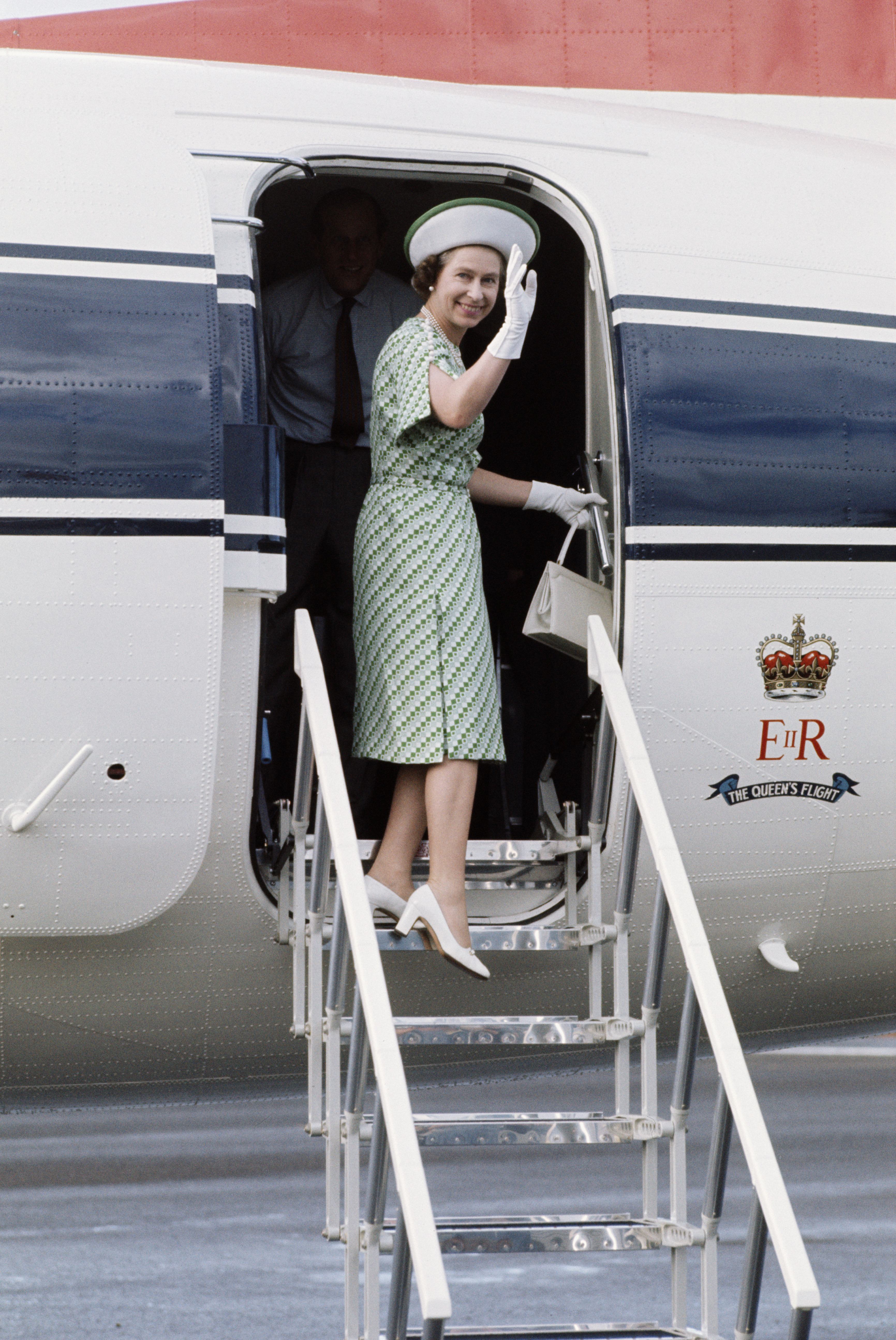 Elizabeth II, wearing a green patterned, short-sleeve dress and a white, wide-brim hat boarding a royal aircraft