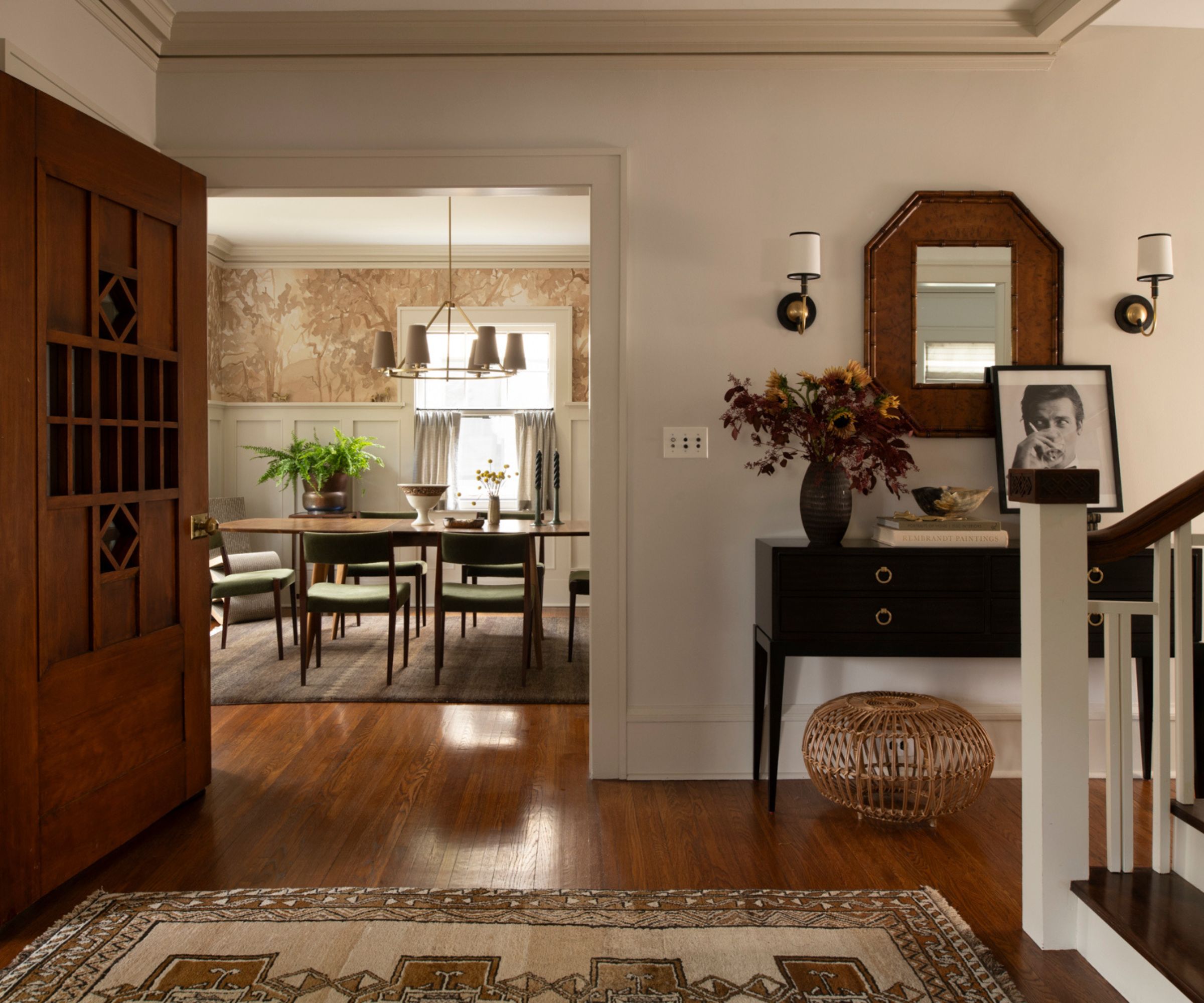 cozy warm entryway decorated for fall with a vintage rug, console table, small rattan ottoman, mirror, sconces, art, and a vase of flowers