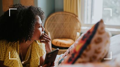 Woman holding phone, looking out of the window while lying relaxed on a bed, scrolling through the best organization apps