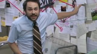 Charlie Day looking stressed out gesturing to a bulletin board filled with paper