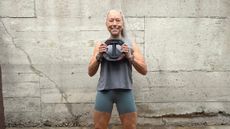 Woman in tank top and cycling shorts stands holding a weight plate smiling at the camera. She stands in front of a gray wall with cracks in it