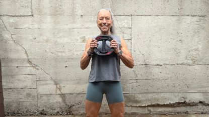 Woman in tank top and cycling shorts stands holding a weight plate smiling at the camera. She stands in front of a gray wall with cracks in it