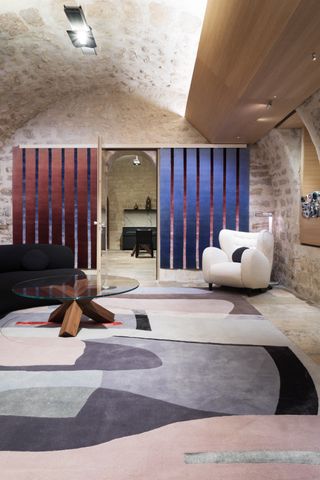 Interior view of Deirdre Dyson’s showroom featuring stone walls and flooring, an arched ceiling and two wine red and blue striped rugs hanging on the wall. There is also a black sofa, a white arm chair with a round black cushion, a multicoloured patterned rug on the floor, a round glass and wooden coffee table and a partial view of another room through an open door
