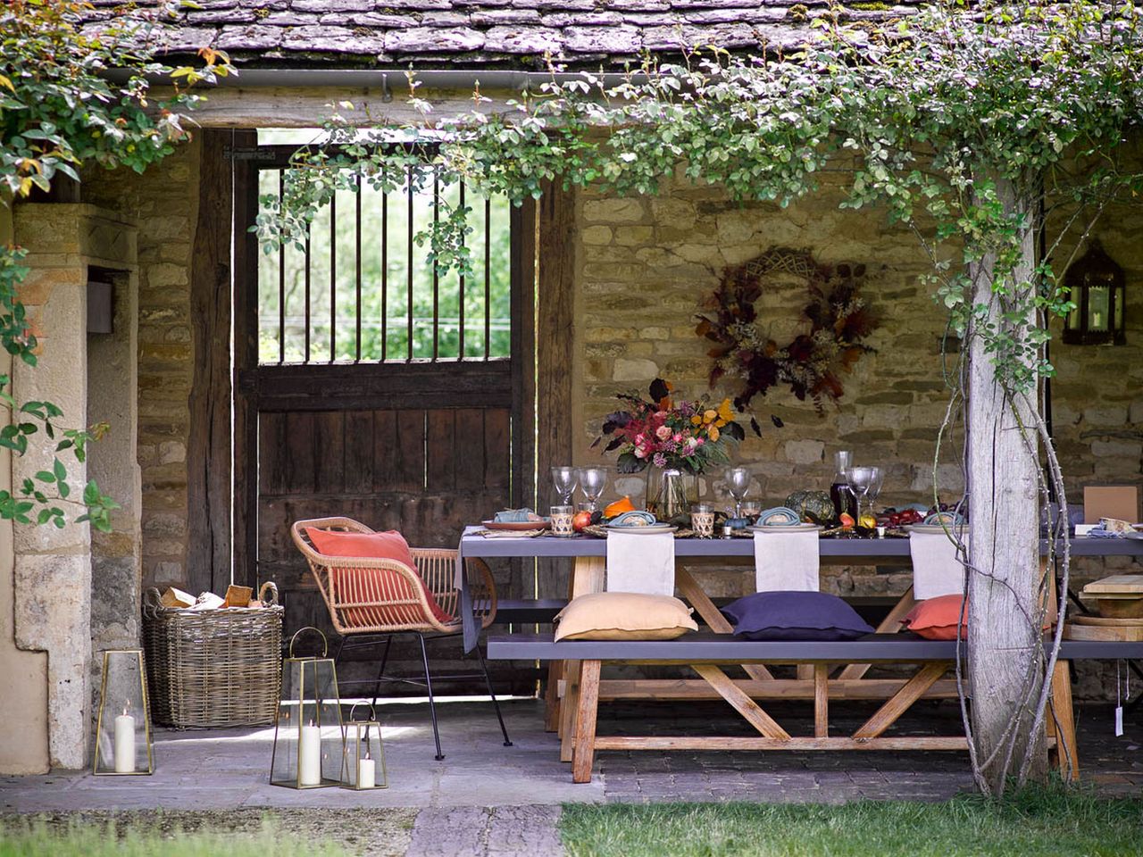 fall table outside with bench and chair seating, vase of fall flowers, fall cushions, pumpkins, wreath on wall, lanterns 