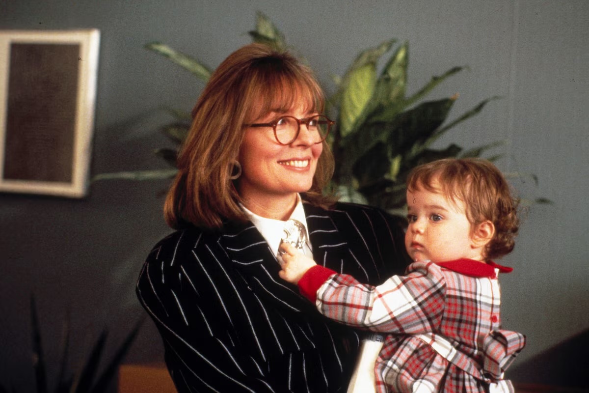 diane keaton wearing glasses and a suit holding a baby in a plaid dress in an office in the movie the baby boom