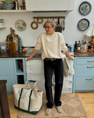 Influencer Lucy Williams poses in her chic London kitchen wearing a cream crewneck sweater, studded Khaite belt, black jeans, and tan suede shoes.