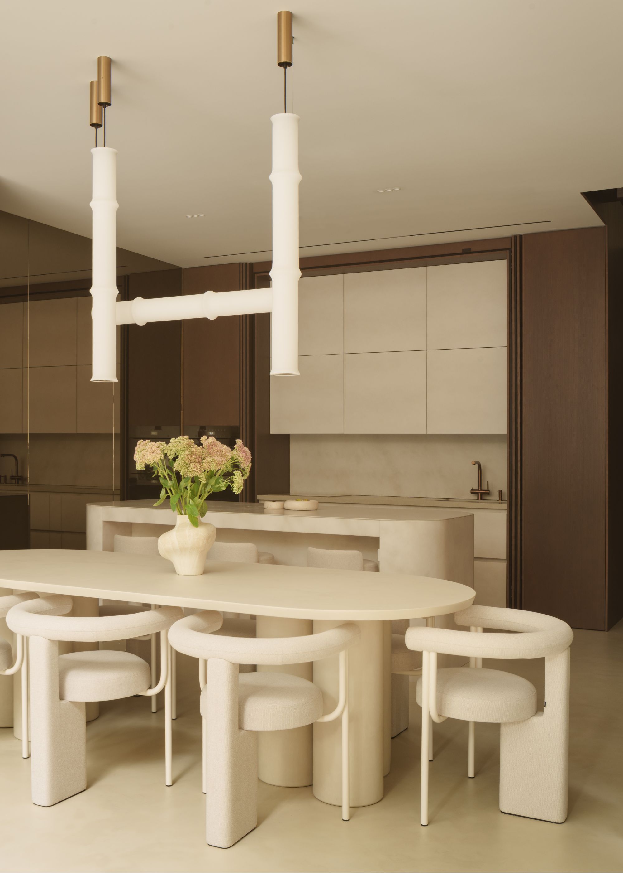 A cream oval-shaped dining table with matching curved cream dining chairs with pendant lighting hanging above, in front of a marble cream kitchen that can be hidden away with sliding timber doors