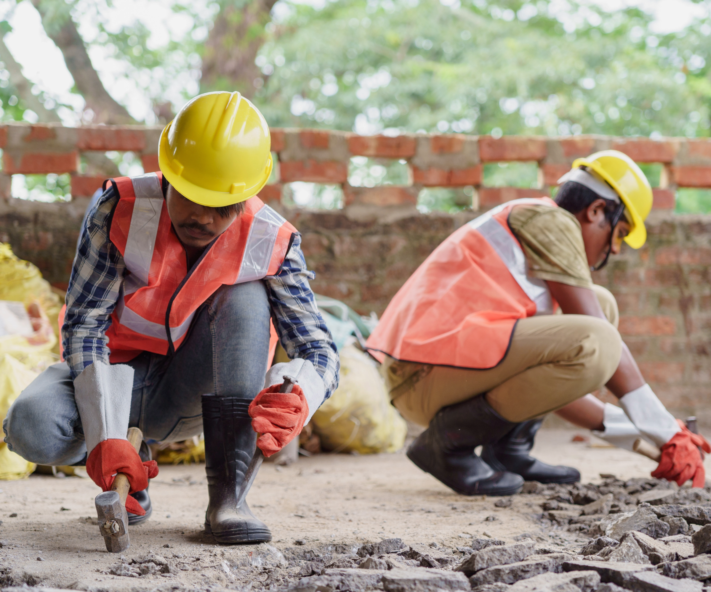 Two builders in hard hats and vests kneeling down and holding a hammer on a construction site