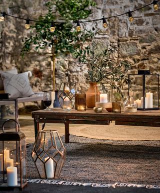 Variety of lanterns on outdoor coffee table and floor, with festoon lighting hung above, and rustic exposed brick wall.