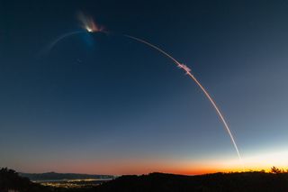 a thin streak of orange light archs through a fading dark sky, originating from the low horizon on the right, where the sky glows orange.